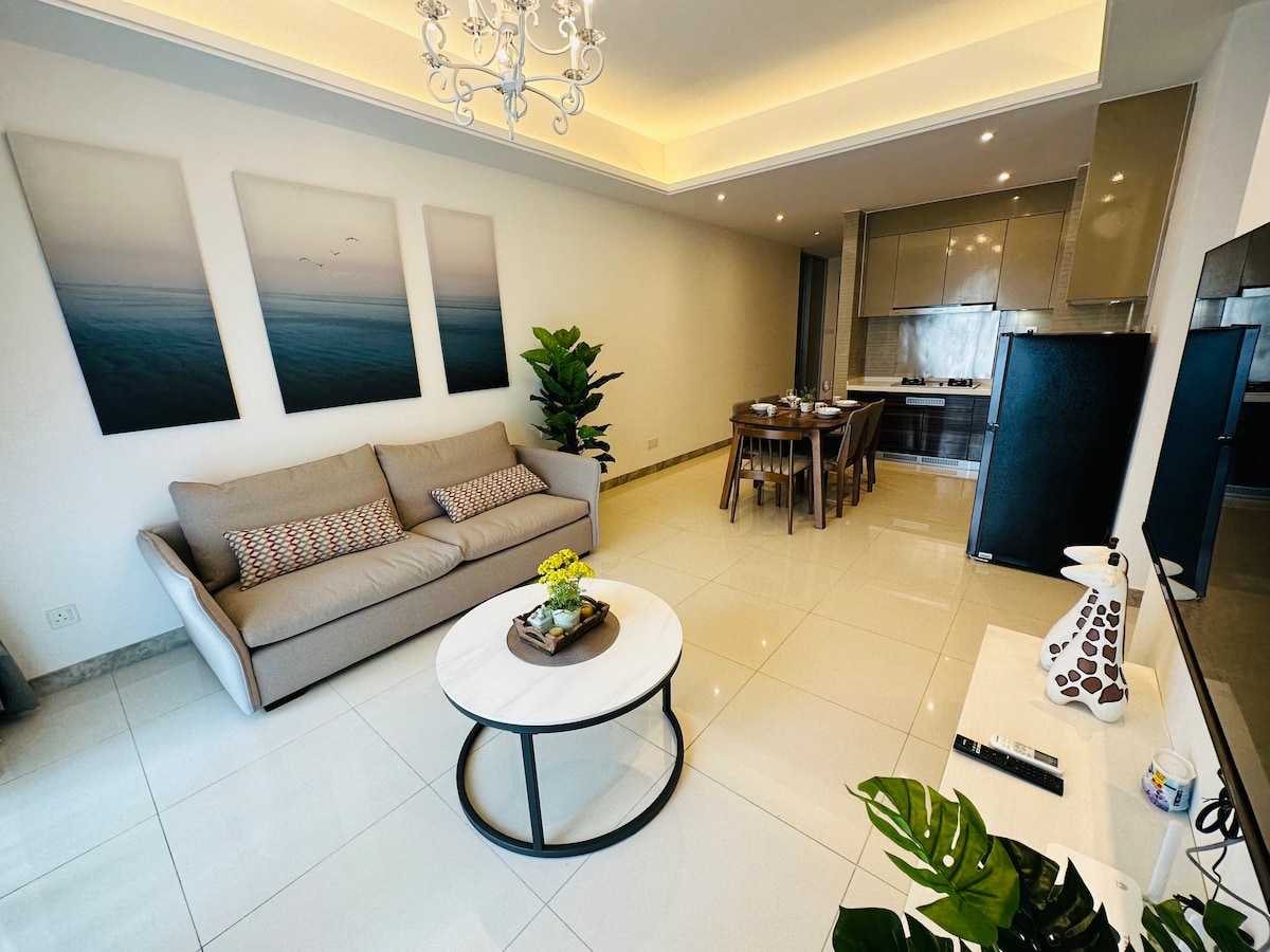 A modern living area features a comfortable sofa across from a circular coffee table. A dining space with wooden chairs and a sleek kitchen is visible in the background. Nature-inspired wall art complements the clean, bright decor, while natural light illuminates the tiled floor.