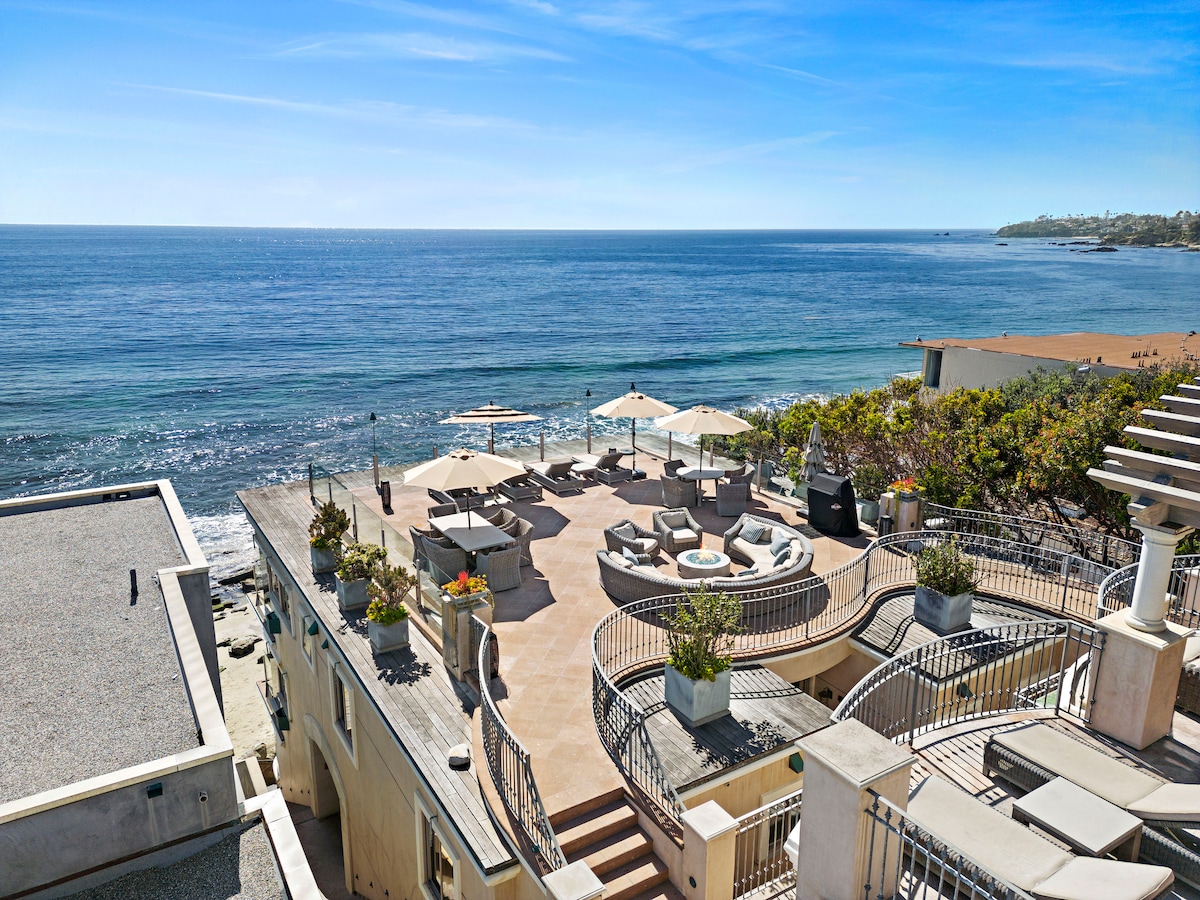 An expansive deck overlooks the ocean, featuring several lounge areas adorned with comfortable seating. Umbrellas provide shade, and potted plants add greenery. The serene coastline and tranquil waters serve as a backdrop, creating a peaceful outdoor retreat.