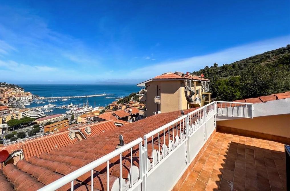 A spacious terrace offers panoramic views of the sea and surrounding hills. Red-tiled rooftops of nearby buildings are visible, along with the marina and blue waters of the gulf. A clear sky enhances the scenic backdrop.
