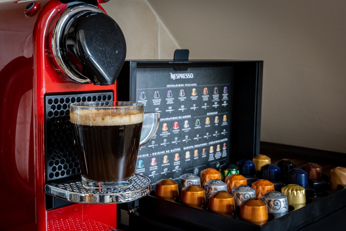 A bright red coffee machine is positioned next to a glass filled with freshly brewed coffee. A variety of colorful coffee capsules are organized in a nearby shelf, showcasing a range of flavors. The smooth surface of the countertop reflects the warm light.