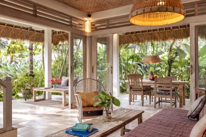 An open living area is surrounded by lush greenery, with large glass doors inviting natural light. Rustic wooden furniture is arranged comfortably, including a patterned sofa and a dining table with chairs. A woven light fixture hangs overhead, adding warmth to the tranquil setting.