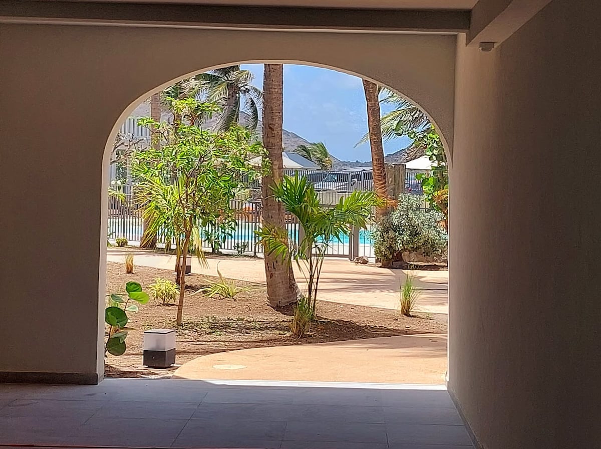 An arched entrance frames a view of palm trees and lush greenery leading to a serene pool area. The outdoor space is well-maintained, featuring sunlight filtering through the foliage and a calm atmosphere conducive to relaxation.