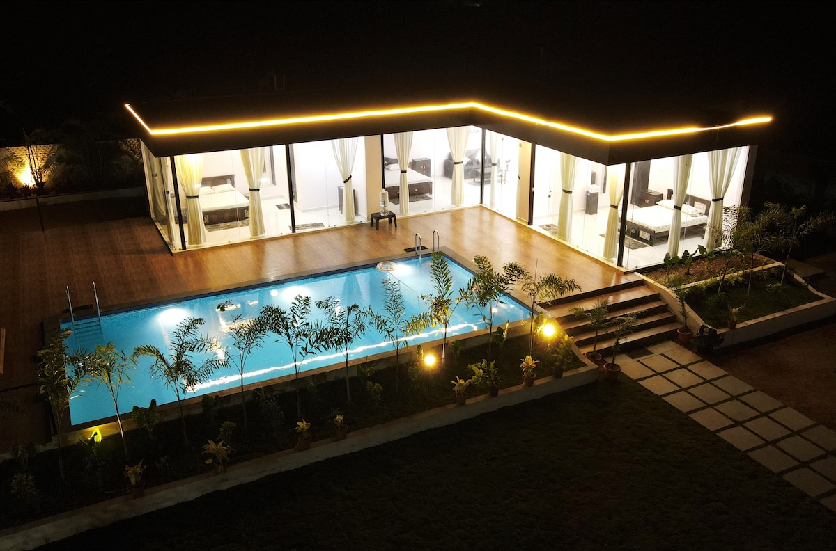 The modern farmhouse is illuminated at night, showcasing a clear swimming pool surrounded by well-maintained greenery. The spacious outdoor area features a patio with seating, while large glass doors provide a view into the tastefully furnished interior.