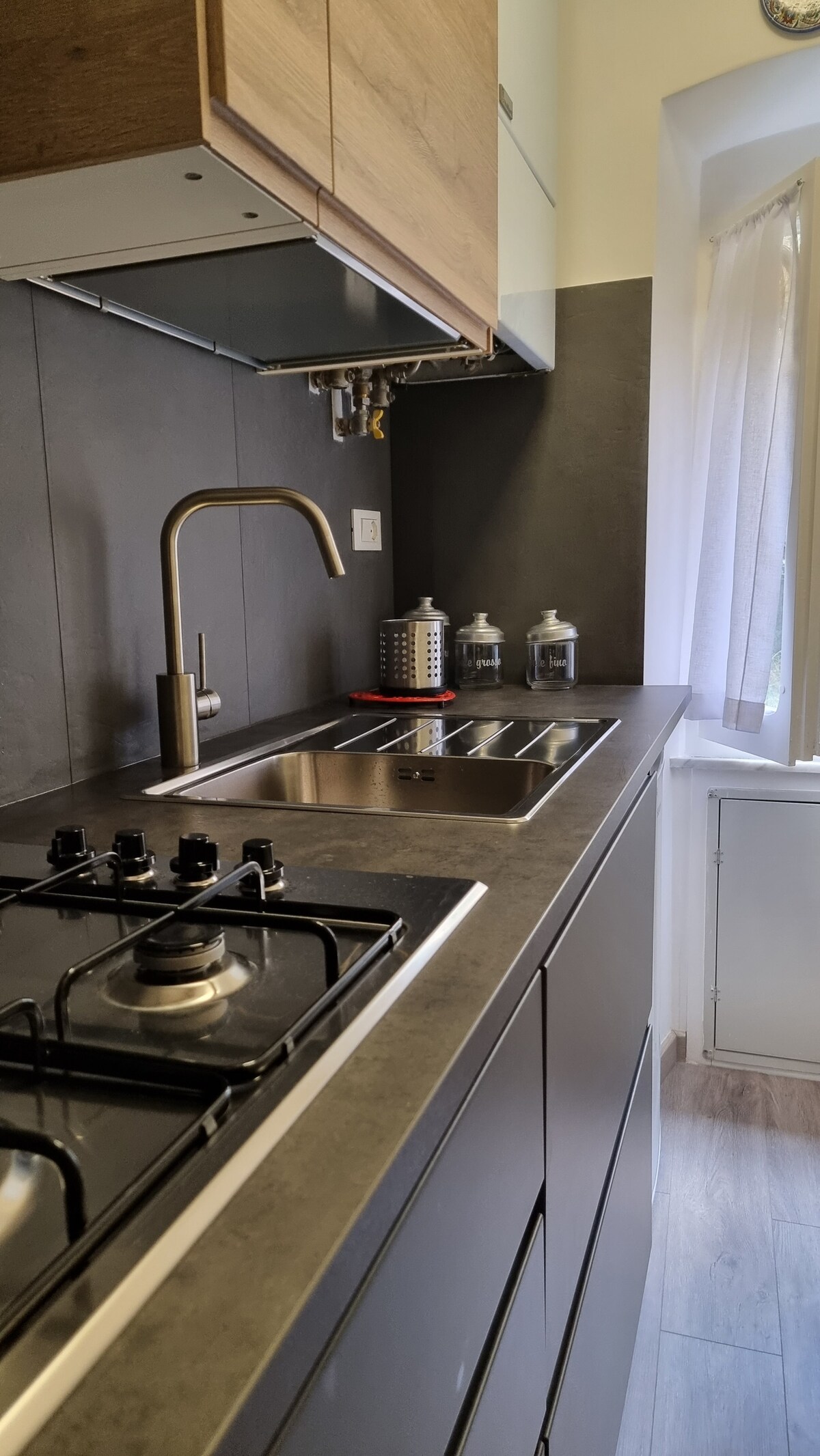 The image showcases a modern kitchen featuring sleek cabinetry with a wood finish. A stainless steel sink and gas stove are prominently displayed against a dark wall. Natural light filters in through a window, illuminating the countertops and nearby decorative elements.
