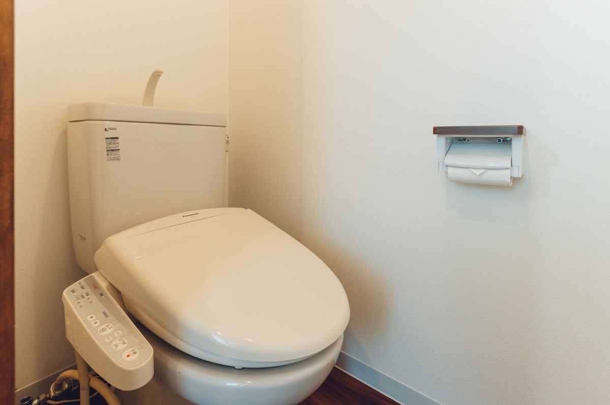 A modern toilet equipped with a Japanese-style bidet is situated against a neutral wall. A small shelf for toilet paper is mounted nearby, and the clean, minimalist design emphasizes functionality within the bathroom space.