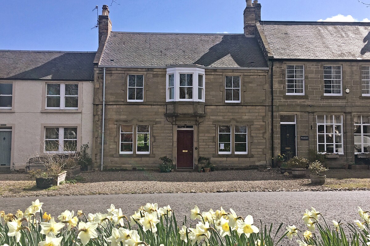 The exterior of a grade-B listed Edwardian townhouse is visible, featuring a traditional stone façade and a prominent bay window. A row of bright daffodils occupies the foreground, drawing attention to the charming village setting with a clear blue sky above.
