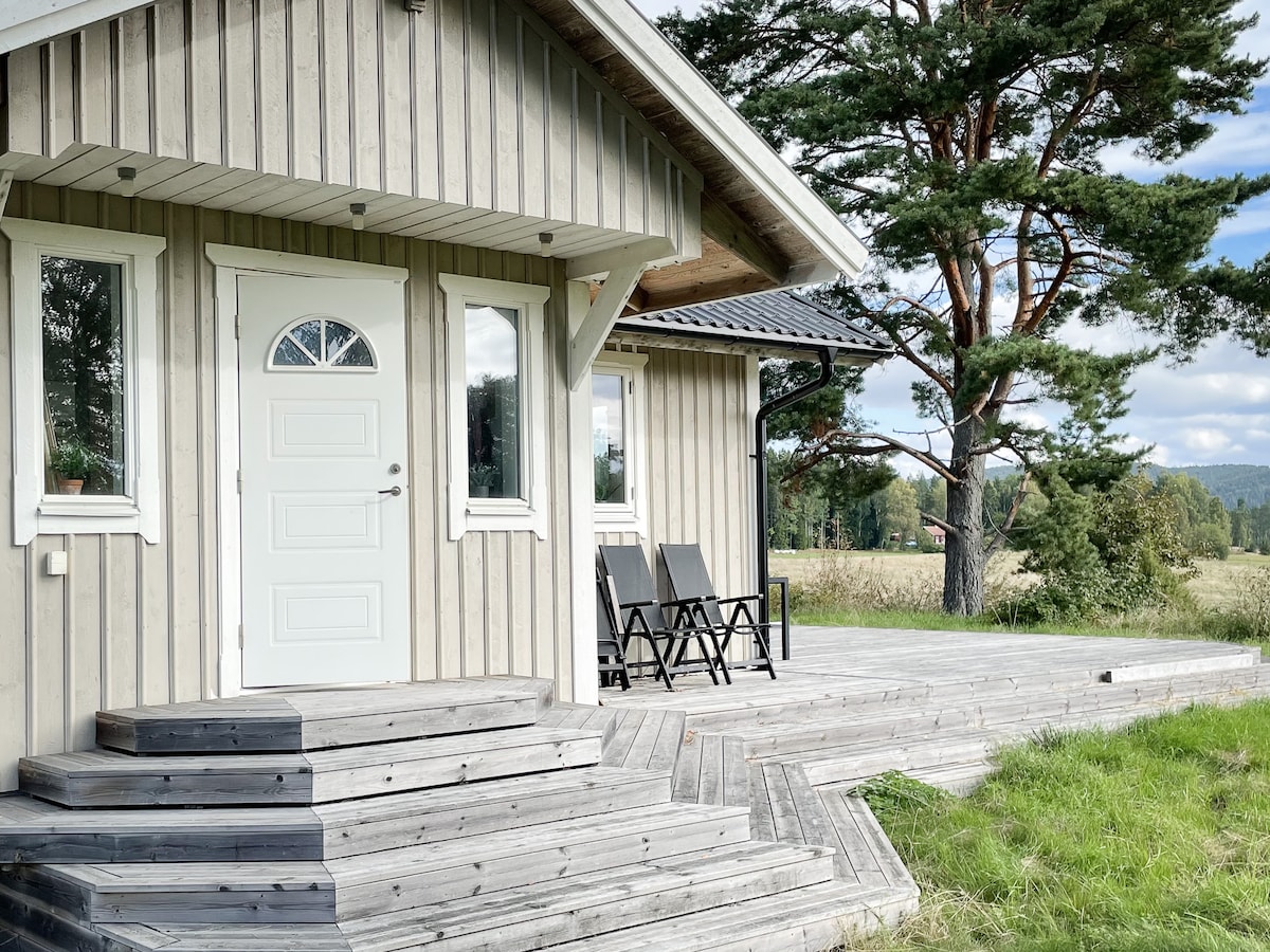 The entrance features a light-colored door framed by a wooden porch, with two reclining chairs positioned on the spacious deck. Surrounding greenery and a backdrop of trees contribute to the peaceful setting, inviting relaxation in the serene natural environment.