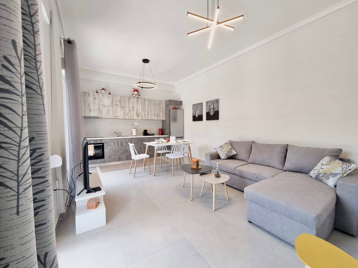 A spacious living and dining area features a light gray sectional sofa, complemented by a modern kitchen with wooden cabinetry. A dining table surrounded by white chairs is visible, alongside a small coffee table. Natural light floods the space through large windows adorned with subtle curtains.