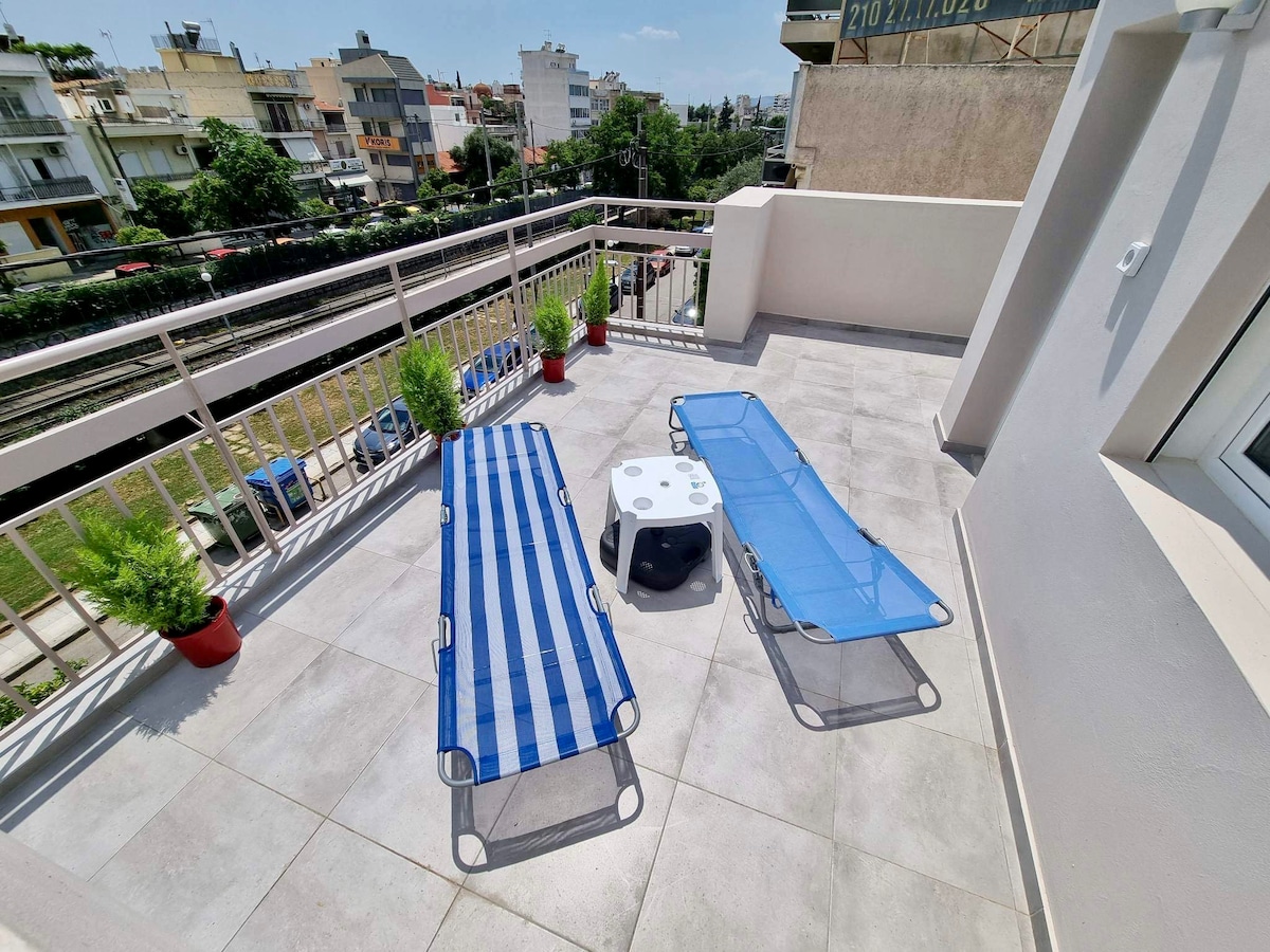 A spacious terrace is shown, featuring two sun loungers with blue fabric and a small white table. Potted plants add a touch of greenery, creating a relaxing outdoor space. The area offers views of the surrounding neighborhood and natural light.