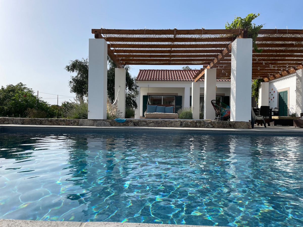 A serene swimming pool reflects the clear sky, offering a view of the spacious outdoor area. The pool is surrounded by white columns and a wooden pergola, providing shade. A comfortable lounging area is visible in the background, enhancing the relaxing atmosphere of the space.