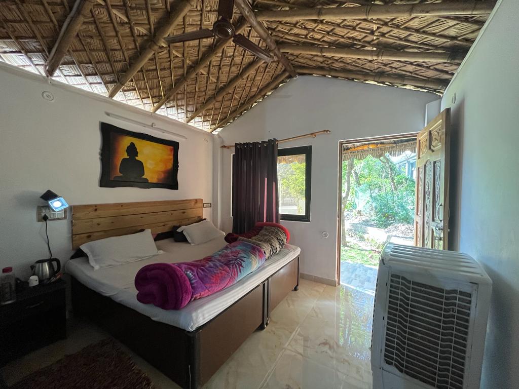 The spacious room features a thatched ceiling and a large bed with colorful bedding. A bedside table holds a lamp, and a kettle is visible on a nearby shelf. Large windows provide views of the lush surroundings, enhancing natural light in the space.