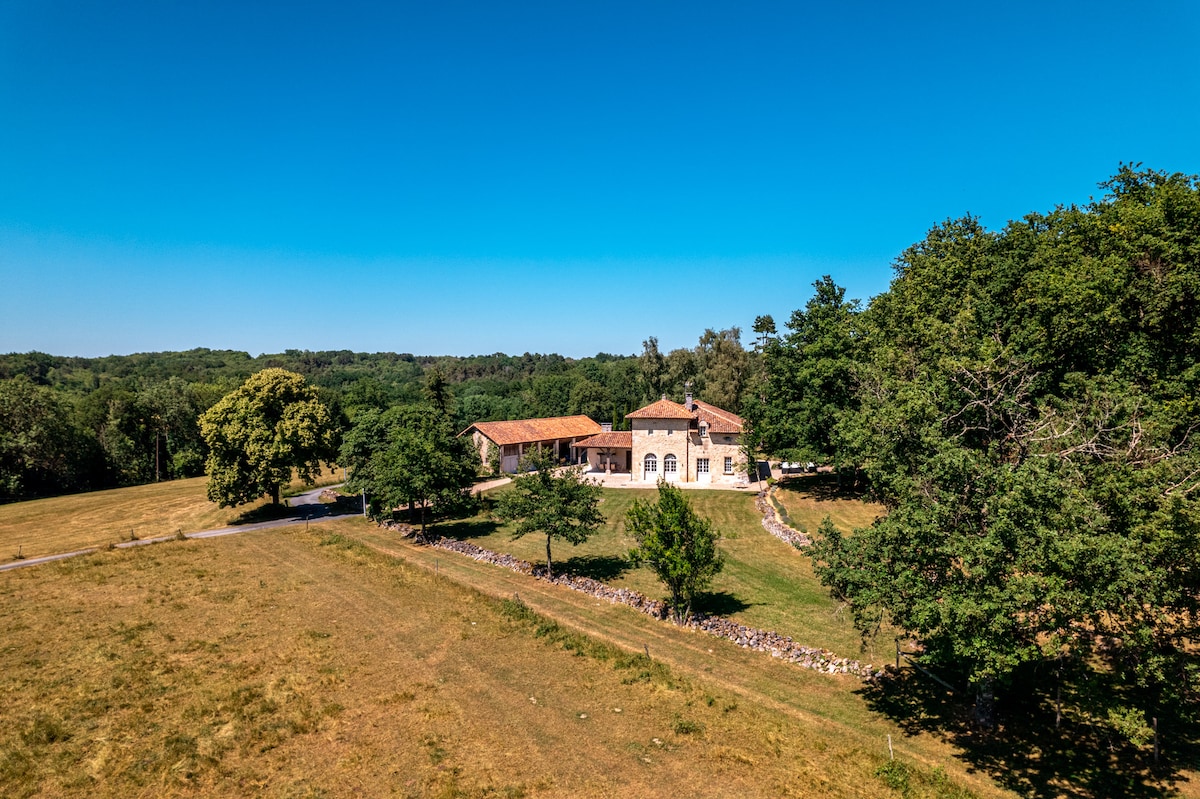 A spacious villa is set amidst a grassy field, surrounded by lush trees and dense woodland. The structure features a combination of rustic stone and terracotta tiles, complemented by a clear blue sky. The tranquil setting offers a sense of privacy and natural beauty.