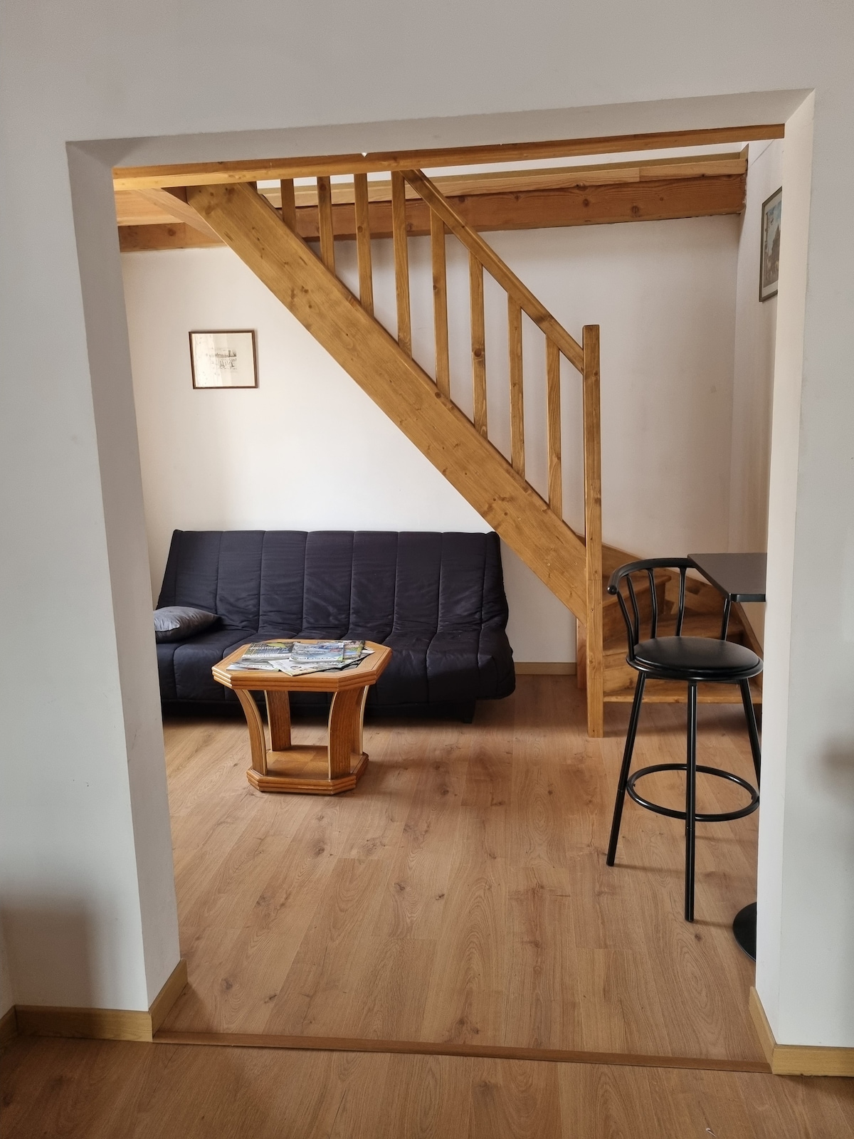An open living area features a black sofa bed, a wooden coffee table, and a high bar stool at a nearby dining area. Natural light fills the space, highlighting the warm wooden beams of the staircase leading to the upper floor.