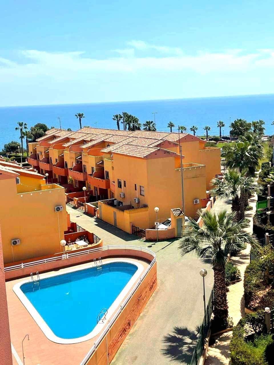 An aerial view showcases a series of vibrant yellow buildings surrounded by lush palm trees and a pool area. The tranquil ocean is visible in the background, enhancing the serene setting. The layout includes pathways leading to the pool, inviting relaxation.