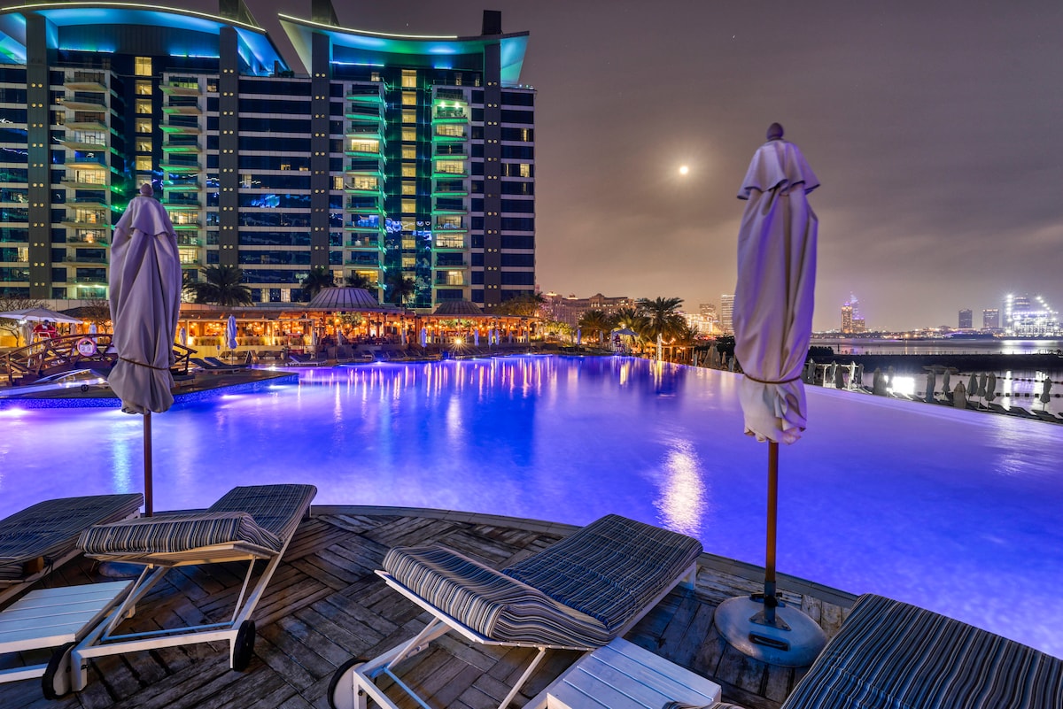 A serene pool area is highlighted by illuminated water reflecting a spectrum of colors. Comfortable lounge chairs are positioned nearby, surrounded by lush palm trees and a vibrant backdrop of the resort buildings. The scene is enhanced by soft moonlight and a clear night sky.