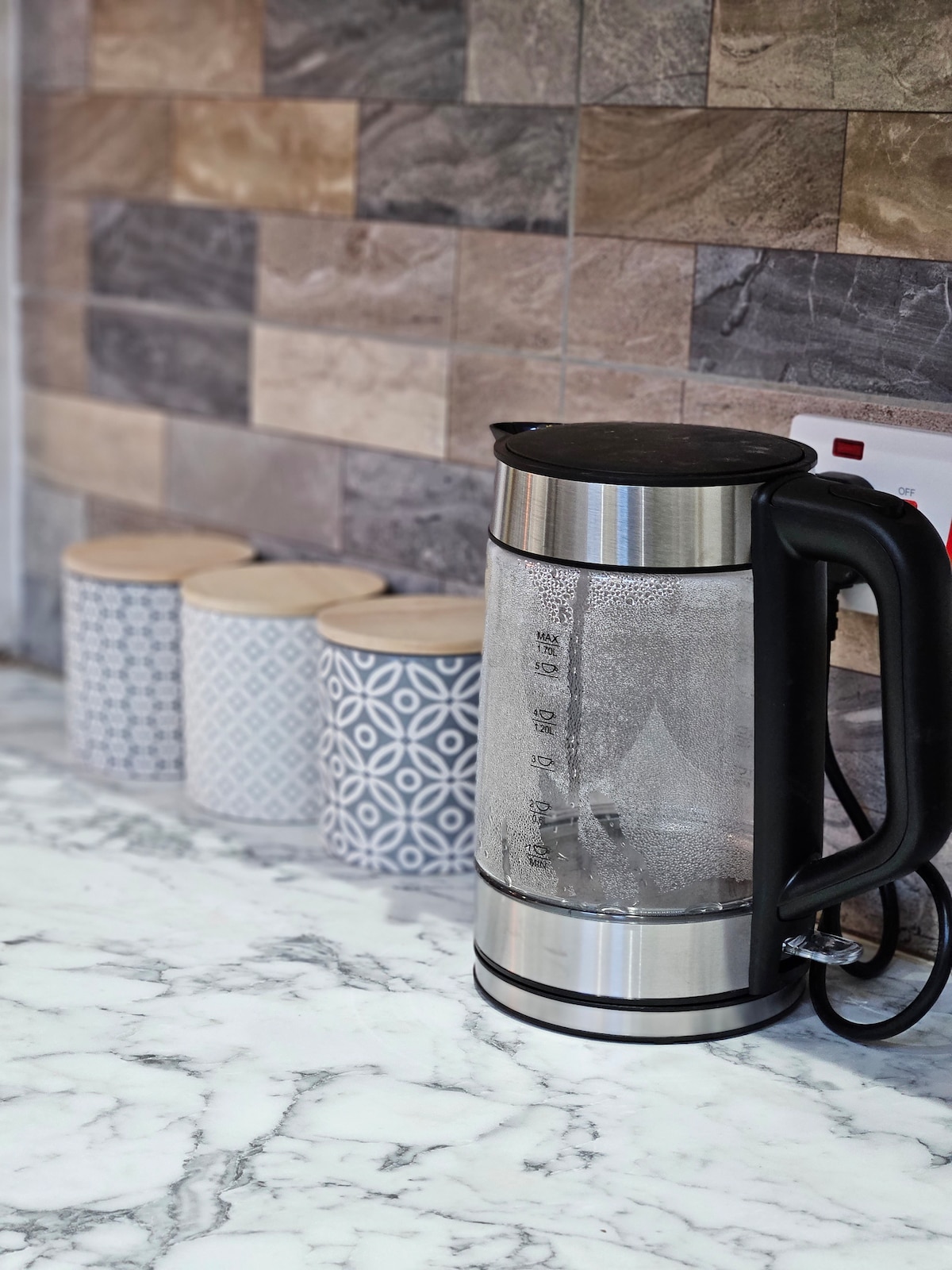 A modern electric kettle rests on a polished marble countertop, with steam rising from its spout. Behind the kettle, three stylish containers with lids are aligned, featuring varying patterns in grey and blue tones against a backdrop of textured stone tiles.