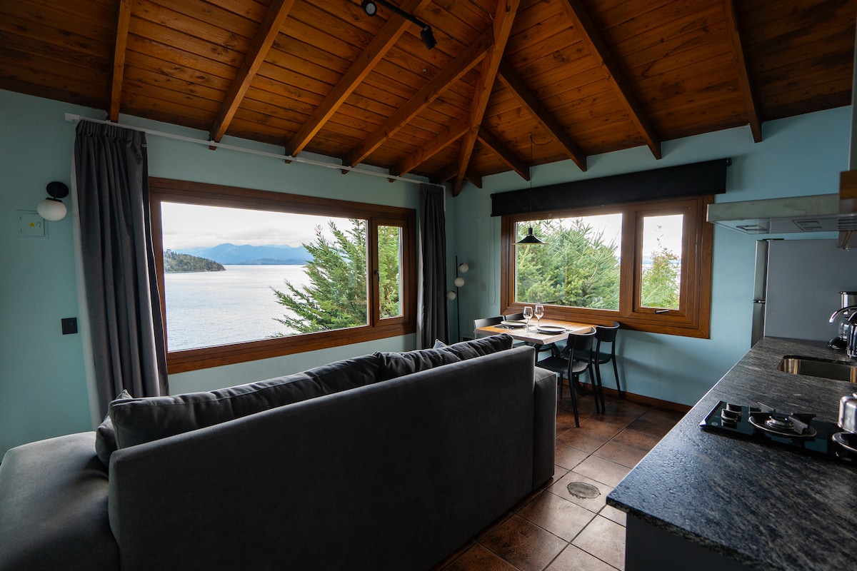 A cozy living area is framed by large windows offering views of the lake and surrounding trees. A comfortable sofa is positioned centrally, accompanied by a dining table set for two. Wooden beams complement the ceiling, adding warmth to the space.