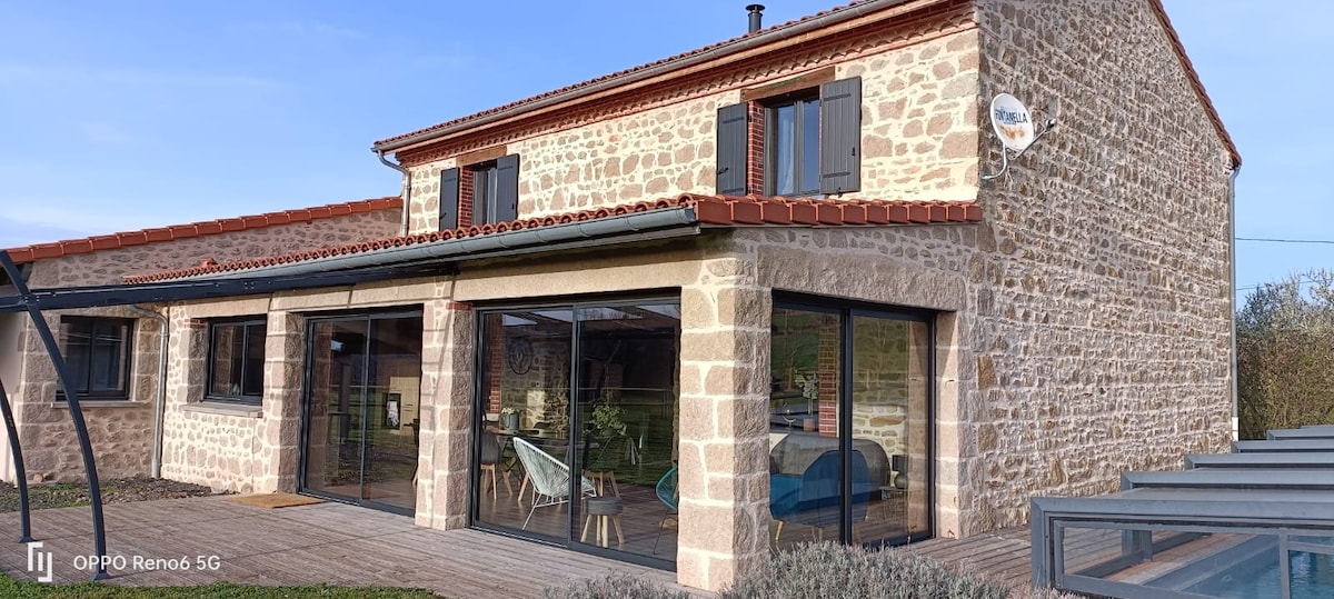 The exterior of a renovated farmhouse is showcased, featuring a stone facade and a tiled roof. Large glass doors provide sunlight to the interior, which is visible through the open space. The surrounding area includes a wooden deck and landscaped greenery.
