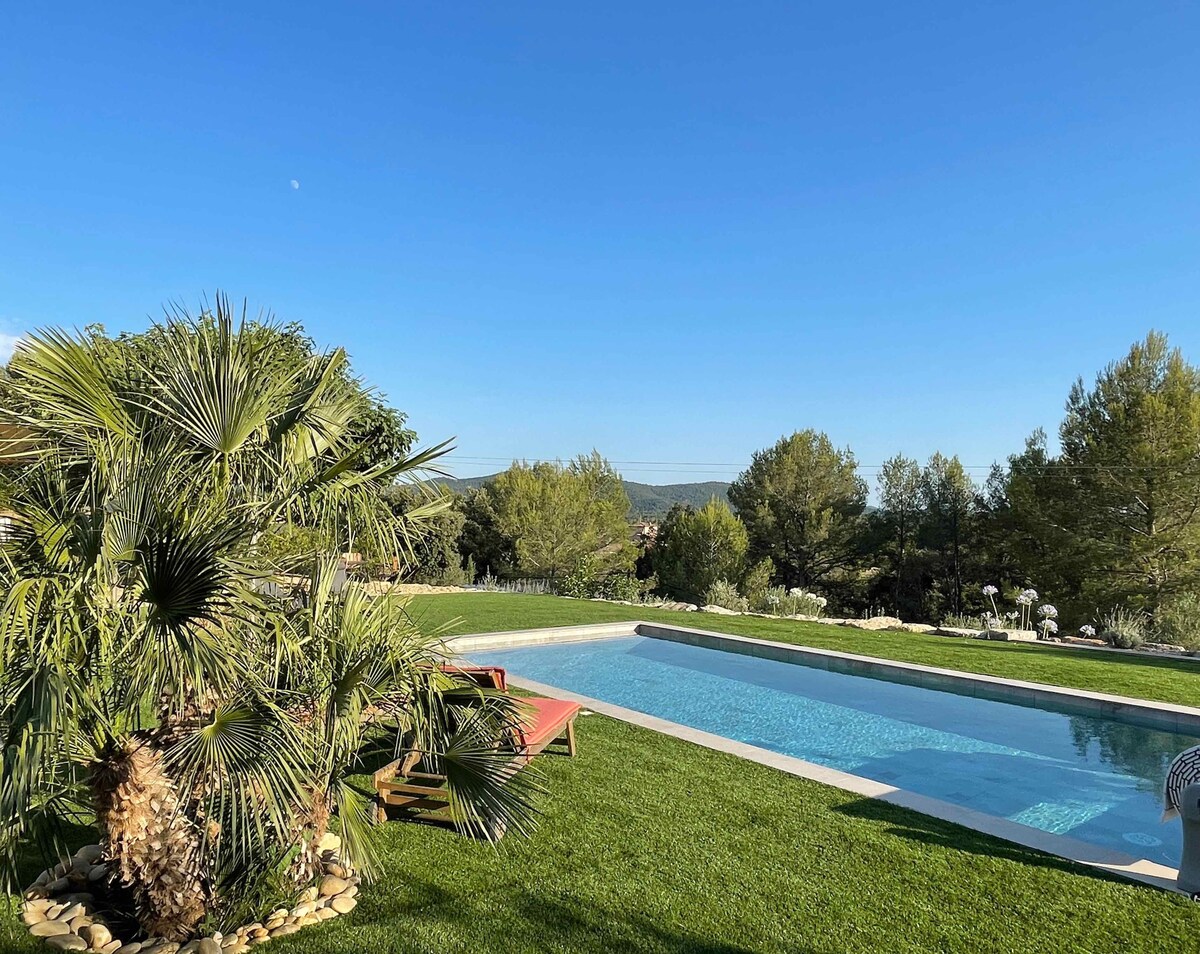 A tranquil swimming pool reflects the clear blue sky, surrounded by lush green grass and a variety of palm trees. Comfortable lounging areas may be seen near the pool, enhancing the serene outdoor space. Views of the nearby hills add to the peaceful ambiance.