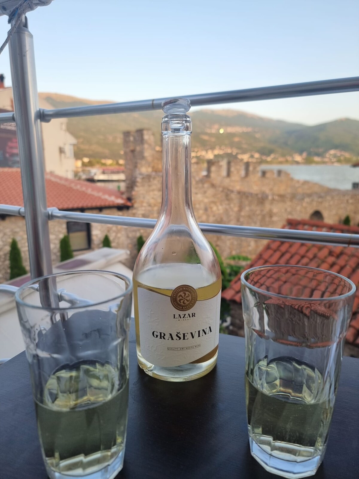 A table is set on a balcony overlooking historical stone walls and rooftops. A bottle of Grasevina wine is positioned at the center, accompanied by two glasses filled with the pale green beverage. The distant mountains and clear sky form a serene backdrop.