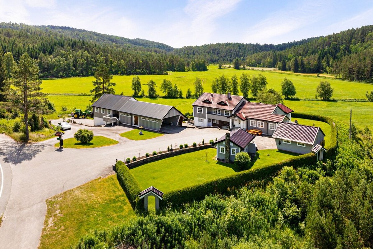 An aerial view of the property showcases a collection of buildings surrounded by manicured lawns and lush greenery. The extensive landscape features rolling fields and a variety of trees, creating a serene atmosphere. A winding pathway leads through the grounds, providing access to each structure.