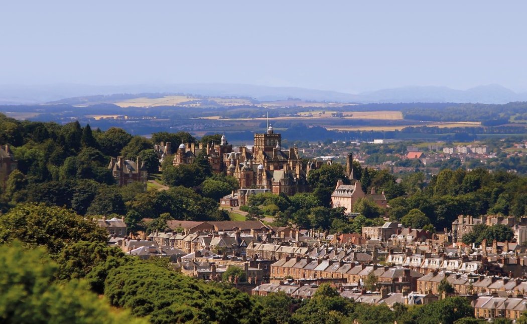 A panoramic view of Edinburgh is presented, showcasing a blend of lush greenery and historic architecture. Rolling hills form the backdrop, with the iconic skyline notable among the treetops. The image captures the tranquil beauty of the city's landscape and its surrounding areas.