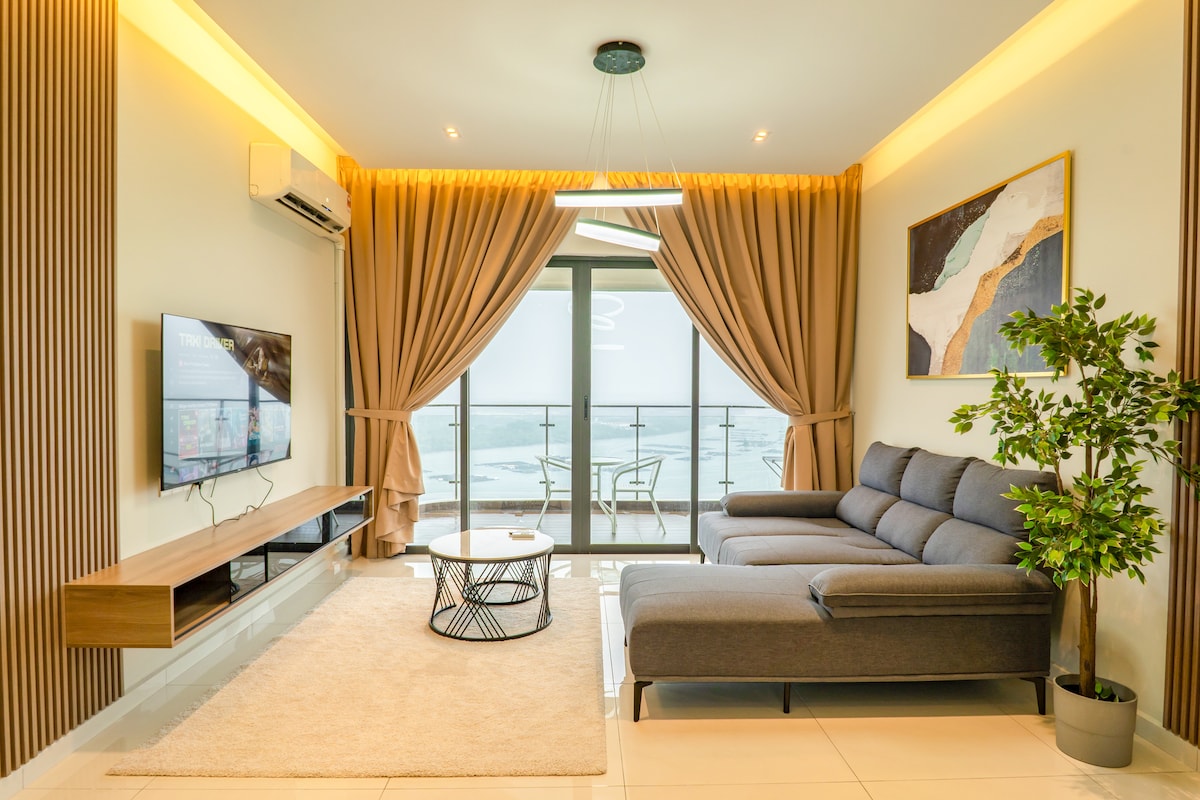 A comfortable living area features a sectional sofa and a decorative coffee table. Floor-to-ceiling windows offer a view of the sea, framed by elegantly draped curtains. A flat-screen television is mounted on the wall, and a potted plant adds a touch of greenery to the space.