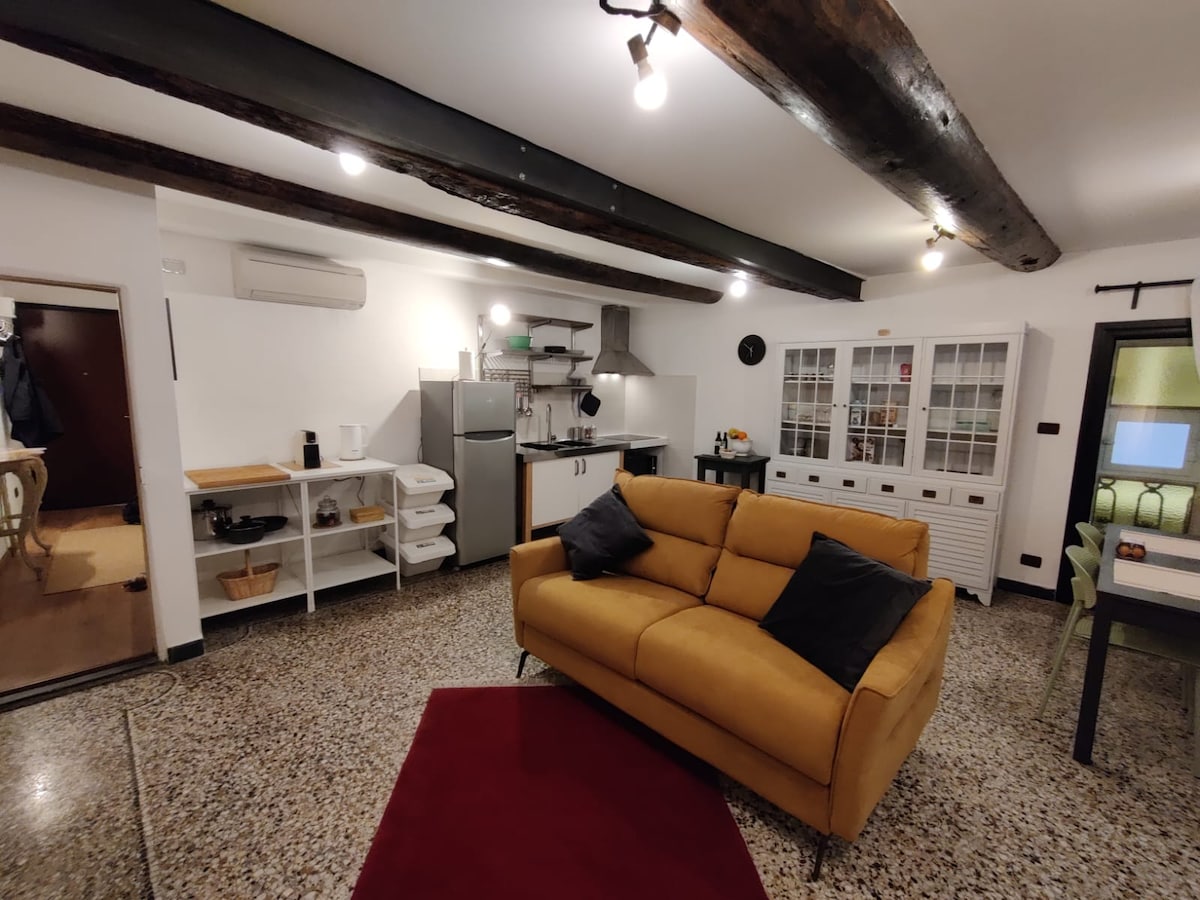 An open living area is featured, showcasing a comfortable yellow sofa with black pillows. The kitchen area is equipped with modern appliances and neatly organized shelves. A red area rug adds a touch of color against the stone floor, and exposed wooden beams highlight the ceiling.