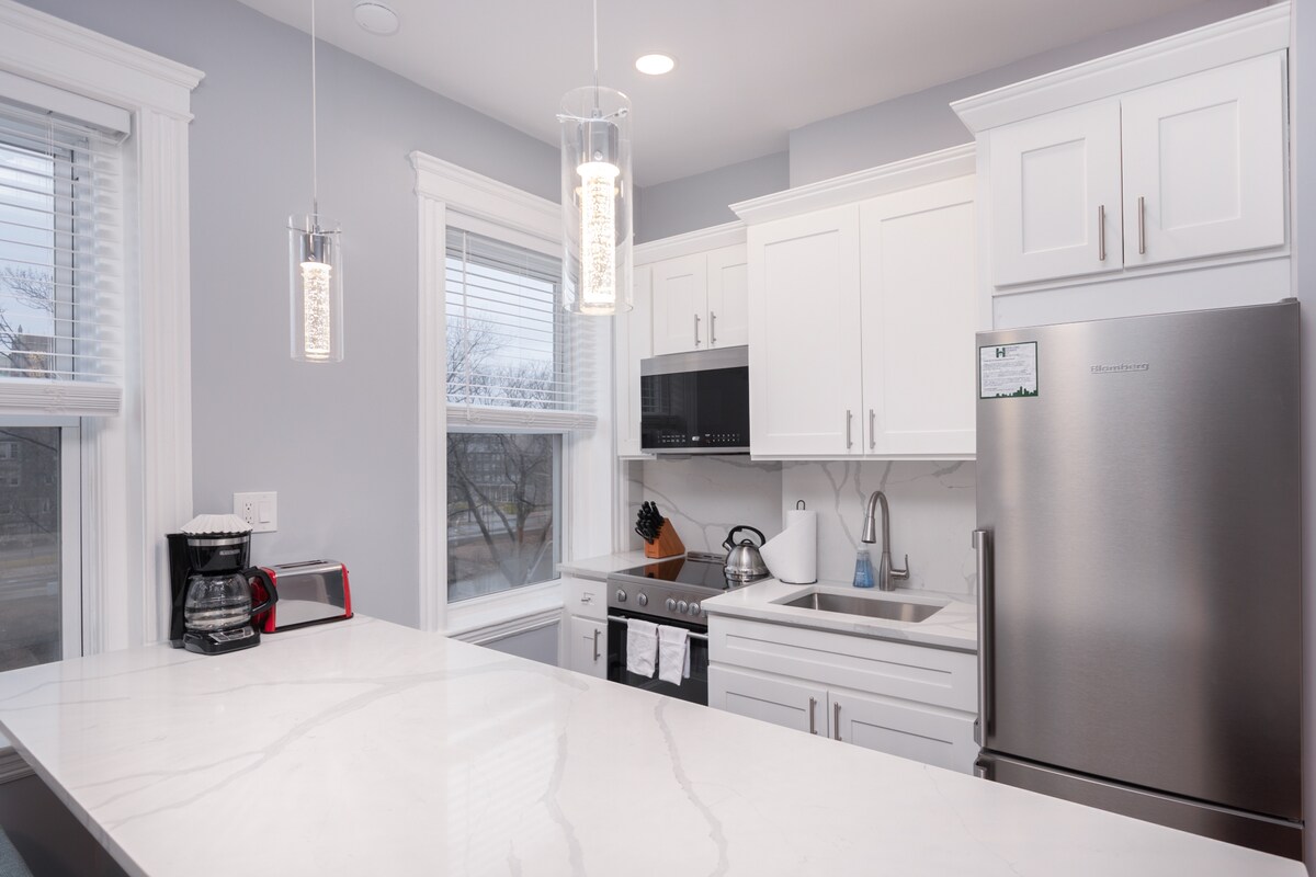 The kitchen features modern white cabinetry with ample storage. Stainless steel appliances, including a refrigerator and microwave, complement the design. A marble countertop provides a spacious area for meal preparation, while large windows bring in natural light, enhancing the overall atmosphere.