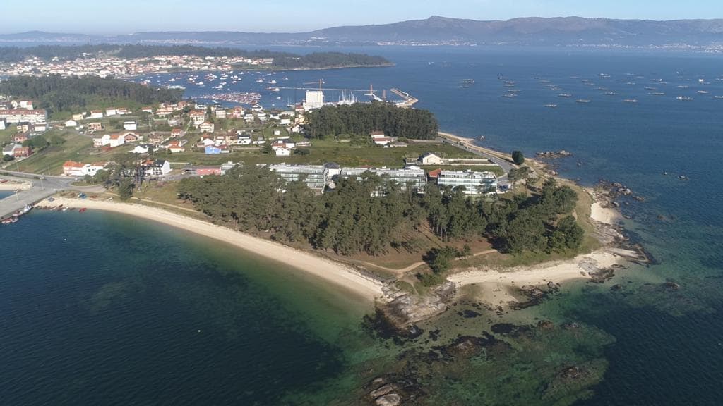 An aerial view showcases the residential area and beaches near the coast. Sandy shores meet the clear blue waters, with boats visible in the distance. Lush greenery surrounds the buildings, enhancing the natural beauty of the Illa de Arousa.