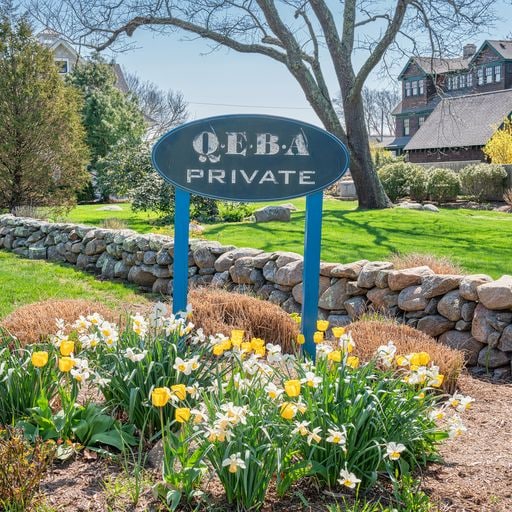 A sign marking the entrance to QUEBA Private is positioned amidst neatly landscaped greenery. Vibrant yellow and white flowers bloom along a stone pathway, contributing to a welcoming appearance. A large tree provides shade, while a traditional house can be seen in the background.
