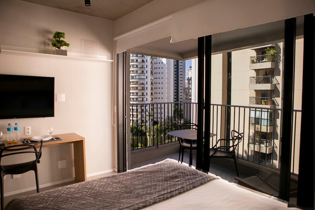 A well-lit studio features large floor-to-ceiling windows overlooking the city. A cozy sleeping area is visible, complemented by a desk and chair setup. A balcony with a small table and chairs invites relaxation while greenery is seen in the surrounding buildings.