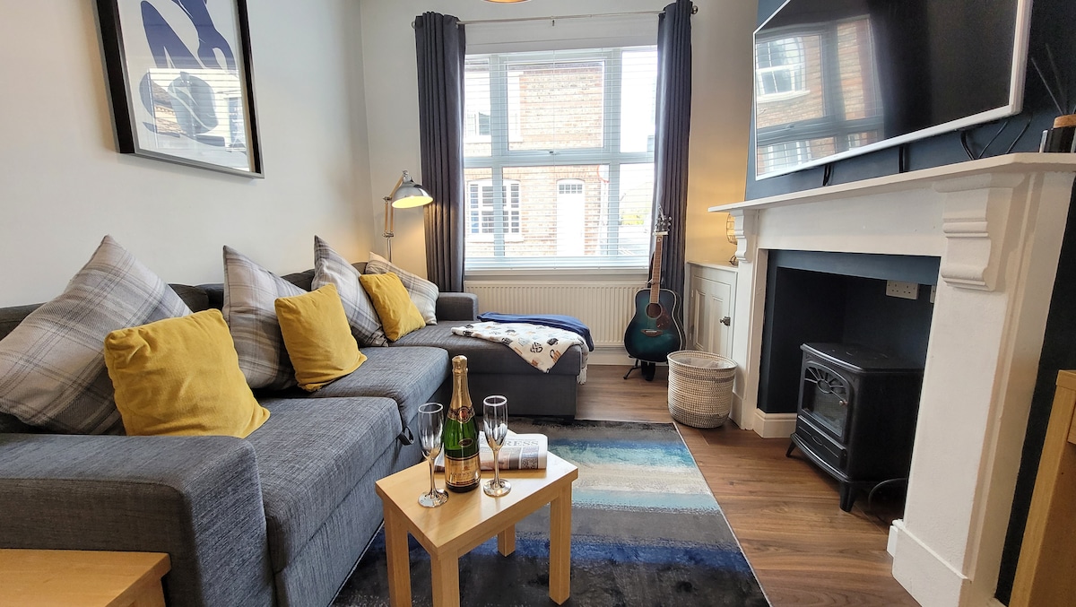 A cozy living space features a gray corner sofa adorned with yellow cushions and a patterned rug. A wooden coffee table holds two champagne glasses and a bottle. A television is mounted above a classic mantelpiece, and natural light enters through the large window.
