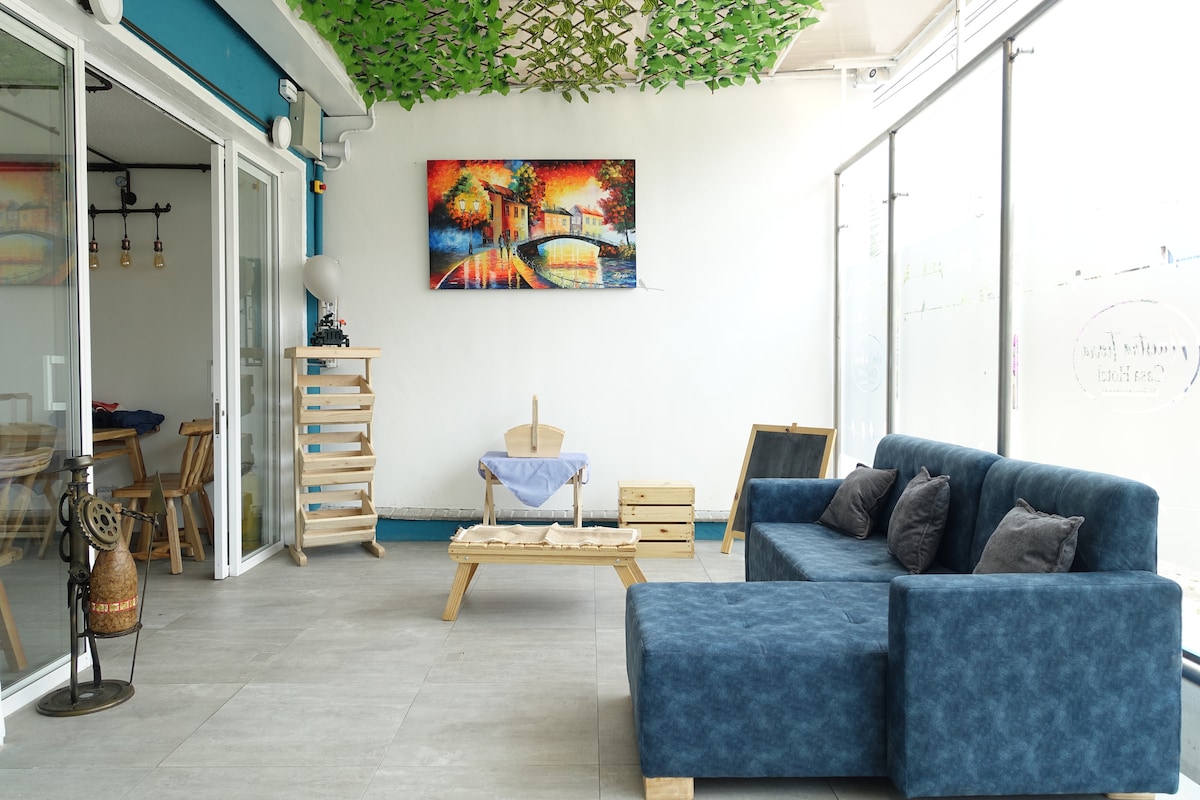 A modern living area is presented with a comfortable blue sofa adorned with cushions. Natural light floods through large glass windows. A wooden coffee table sits at the center, surrounded by simple wooden furniture. A vibrant artwork decorates the wall, enhancing the space.
