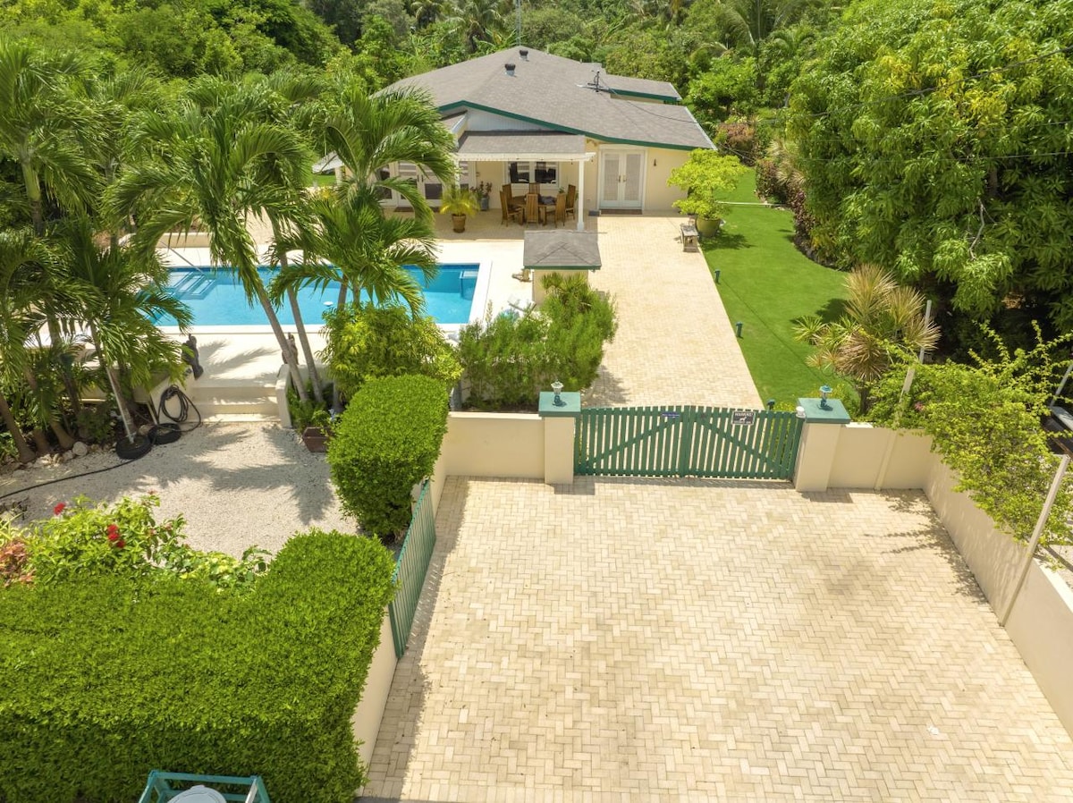 An aerial view captures the home amidst its lush surroundings. The inviting saltwater pool is visible alongside the patio area with seating. A meticulously maintained garden with tropical greenery enhances the serene outdoor environment, while a gated entrance leads to the driveway.