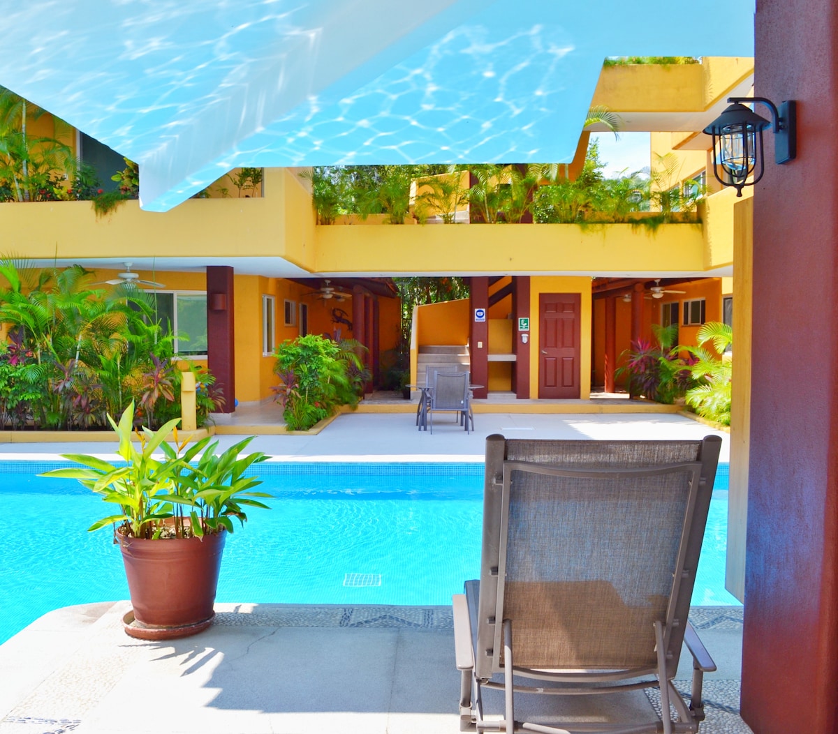 A spacious shared pool area is framed by vibrant greenery and terraces. Comfortable seating is provided beside the pool, allowing relaxation while enjoying the soothing water. The surrounding environment features colorful plants, enhancing the tranquil outdoor experience.