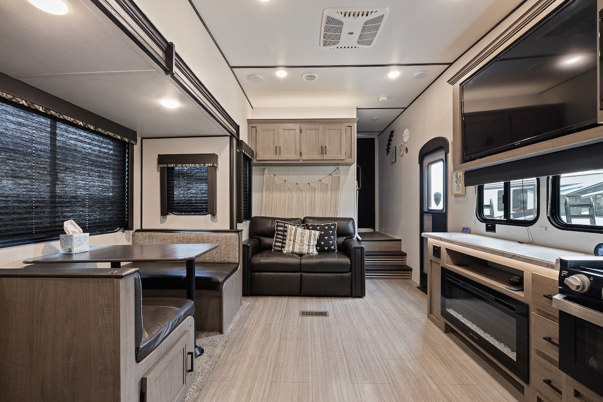 An open living space features a dark leather sofa and a wooden dining table with chairs. Black window shades allow for privacy, while natural light filters in. A mounted television and decorative wall accents add functional and stylistic elements to the modern interior.