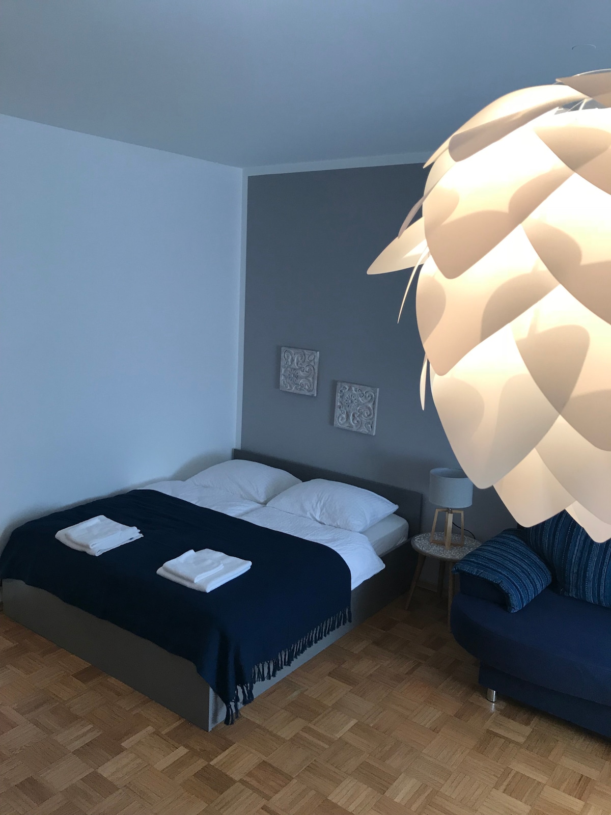 A cozy sleeping area features a spacious bed dressed in white linens, adorned with a navy blue throw blanket. Two towels are neatly placed at the foot of the bed. A modern fixture hangs overhead, while a comfortable chair is positioned nearby.