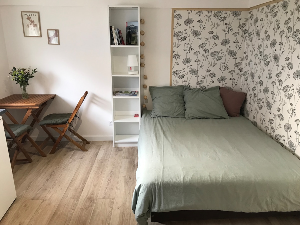 A bedding area features a spacious bed dressed in soft green linens, accompanied by a decorative accent pillow. A small wooden table with chairs is positioned nearby, while a white shelf displays books and a lamp, adding to the room's practical layout.