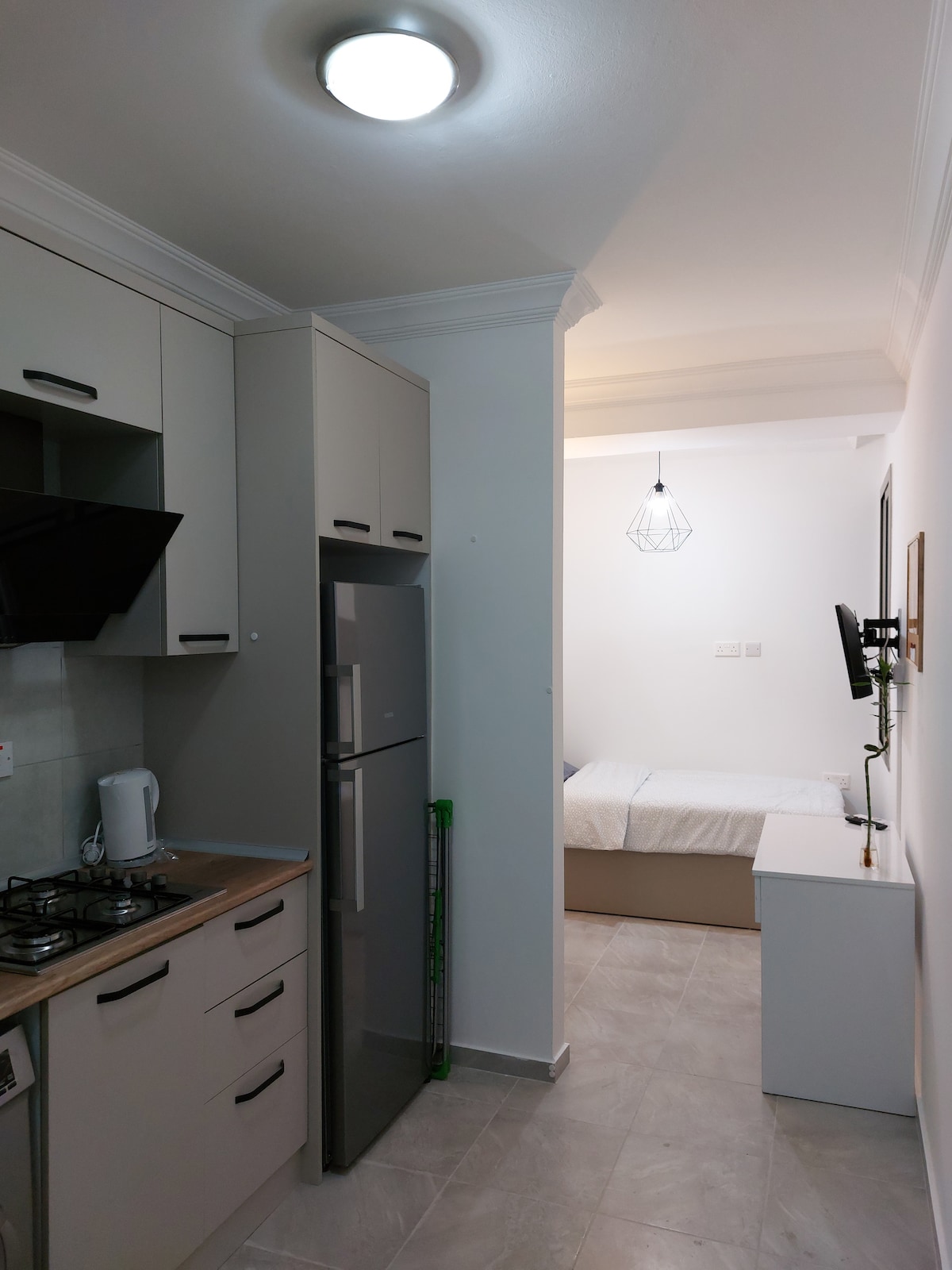 A compact kitchen area is visible, featuring modern cabinetry, a stove, and a refrigerator. A soft light fixture hangs above, illuminating the space. In the background, a cozy sleeping area is discernible with a neatly made bed and a small bedside table.