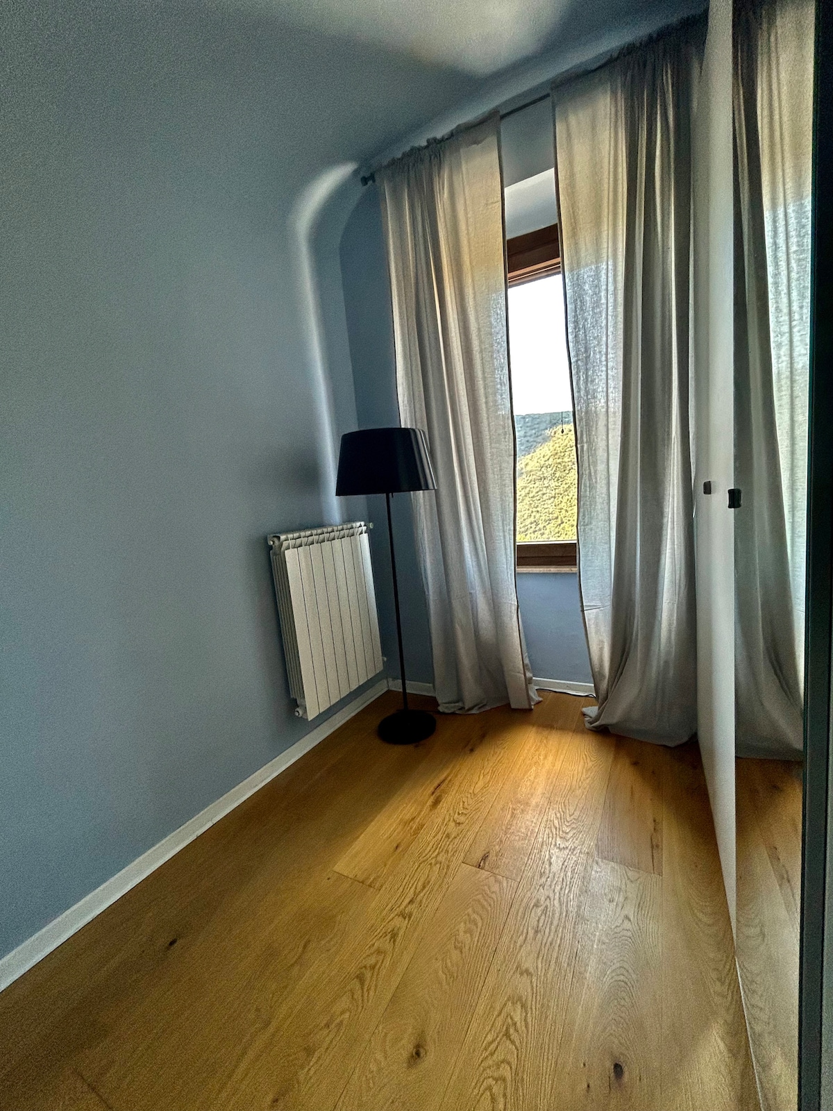 A corner of a room features a window dressed with flowing curtains that softly diffuse natural light. A tall, slender lamp stands beside a modern radiator on a wooden floor, adding warmth to the space. The walls are painted in light blue, creating a serene atmosphere.