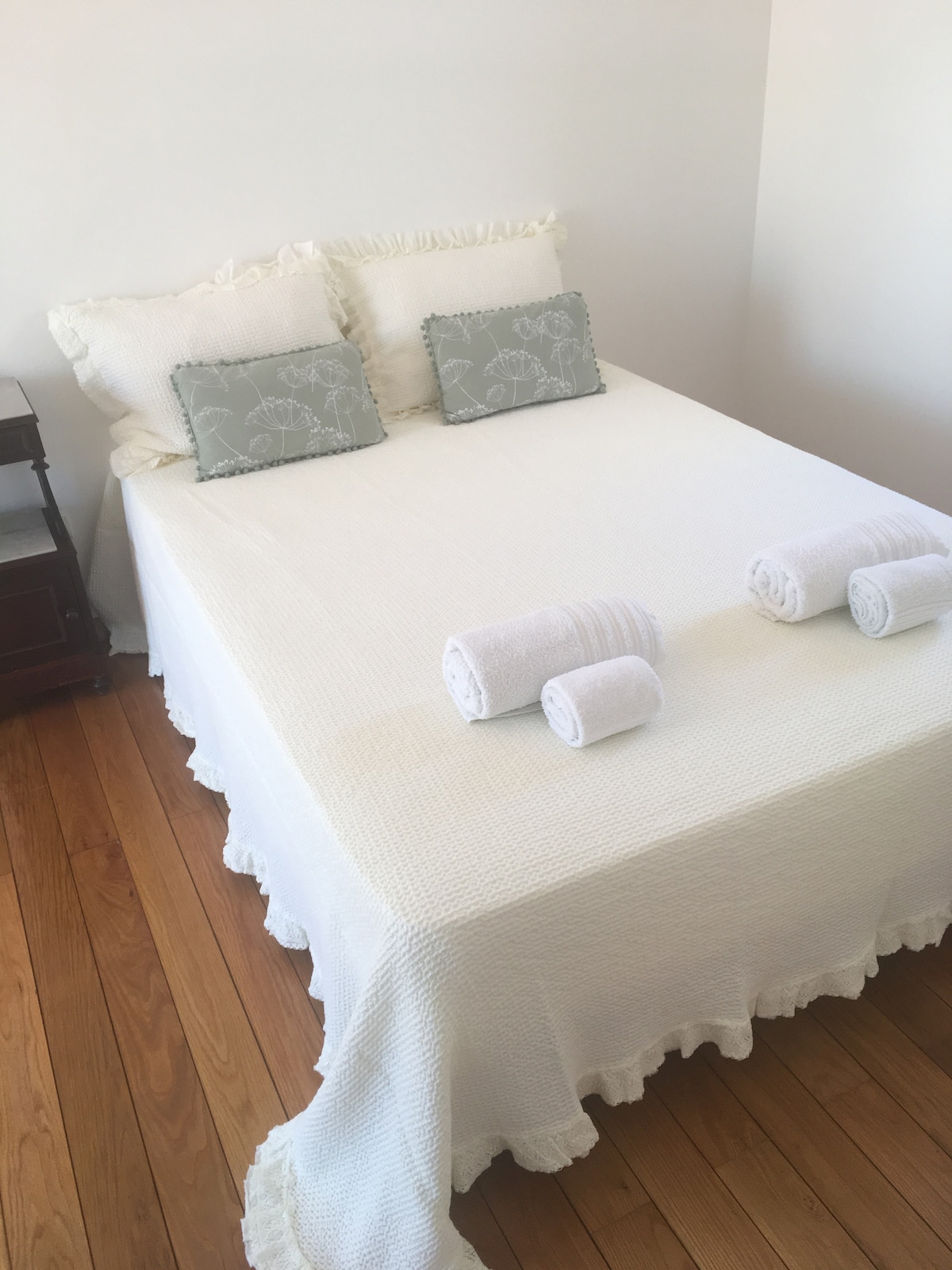 A bed with a textured white quilt is displayed, complemented by two decorative pillows with patterns. Freshly rolled towels are positioned neatly at the foot of the bed. Natural light illuminates the warm wooden flooring, enhancing the overall simplicity of the sleeping space.