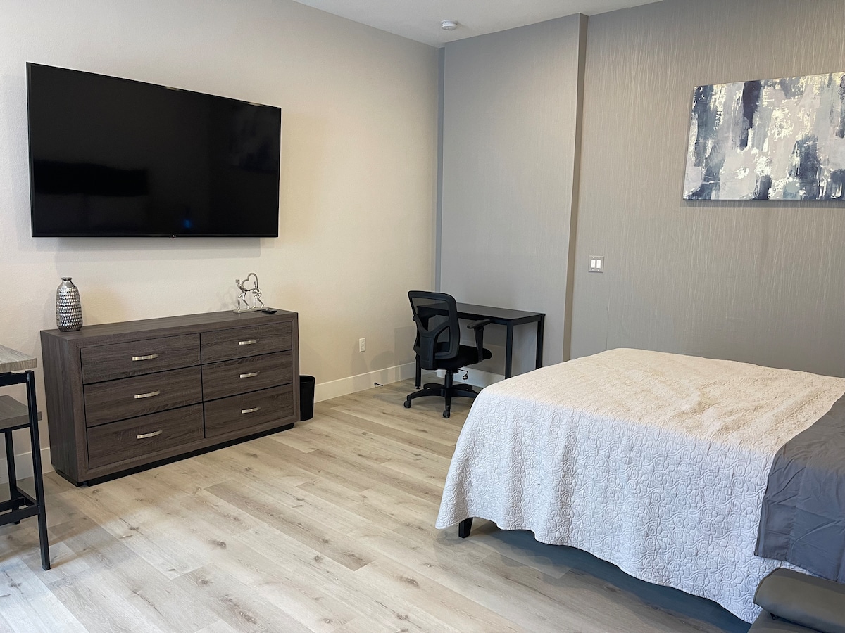 A spacious room is presented with light wood flooring and neutral colored walls. A queen mattress is dressed in a light coverlet, while a television is mounted on the wall across from a desk and chair. A dresser with multiple drawers offers additional storage.