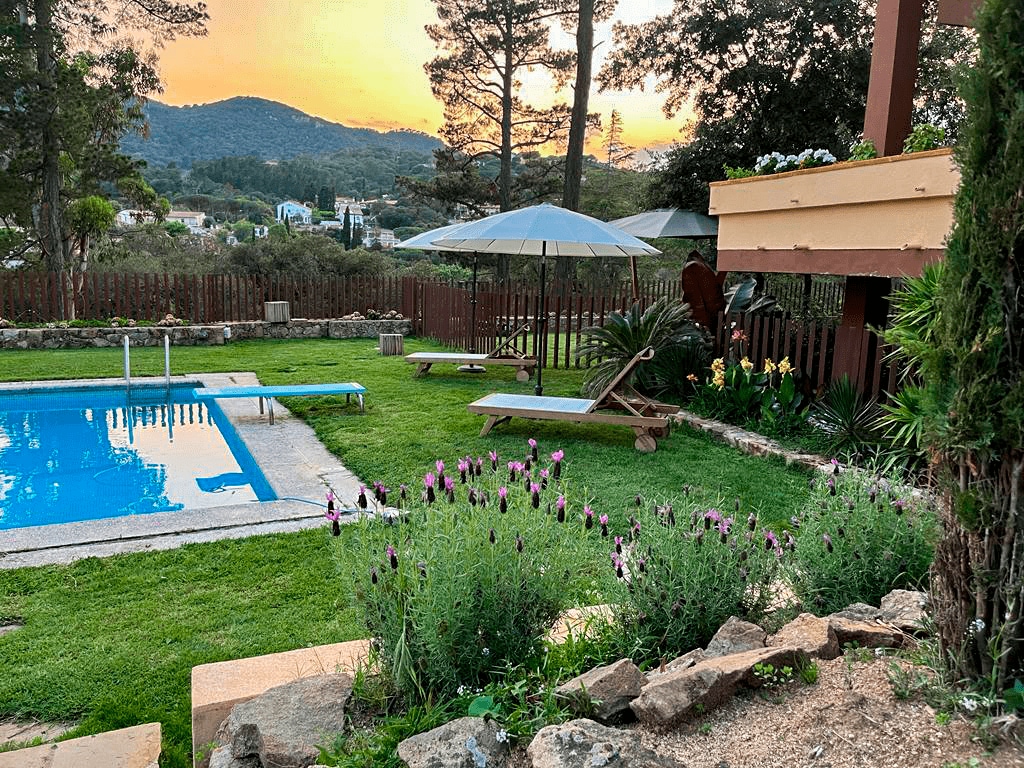 A tranquil garden area features a swimming pool surrounded by well-kept grass and vibrant flowers. A shaded umbrella provides comfort beside a wooden seating area. The scene is set against a backdrop of mountainous terrain and a sunset sky, contributing to a serene outdoor atmosphere.