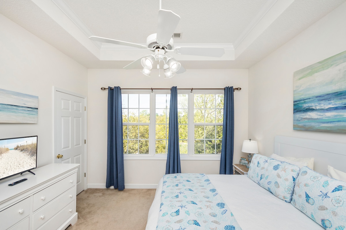 A bright bedroom features a spacious bed adorned with blue and white bedding. Natural light streams through two large windows, framed by blue curtains. A ceiling fan aids air circulation, and a 32” smart TV is placed on a dresser opposite the bed. Coastal-themed artwork decorates the walls.