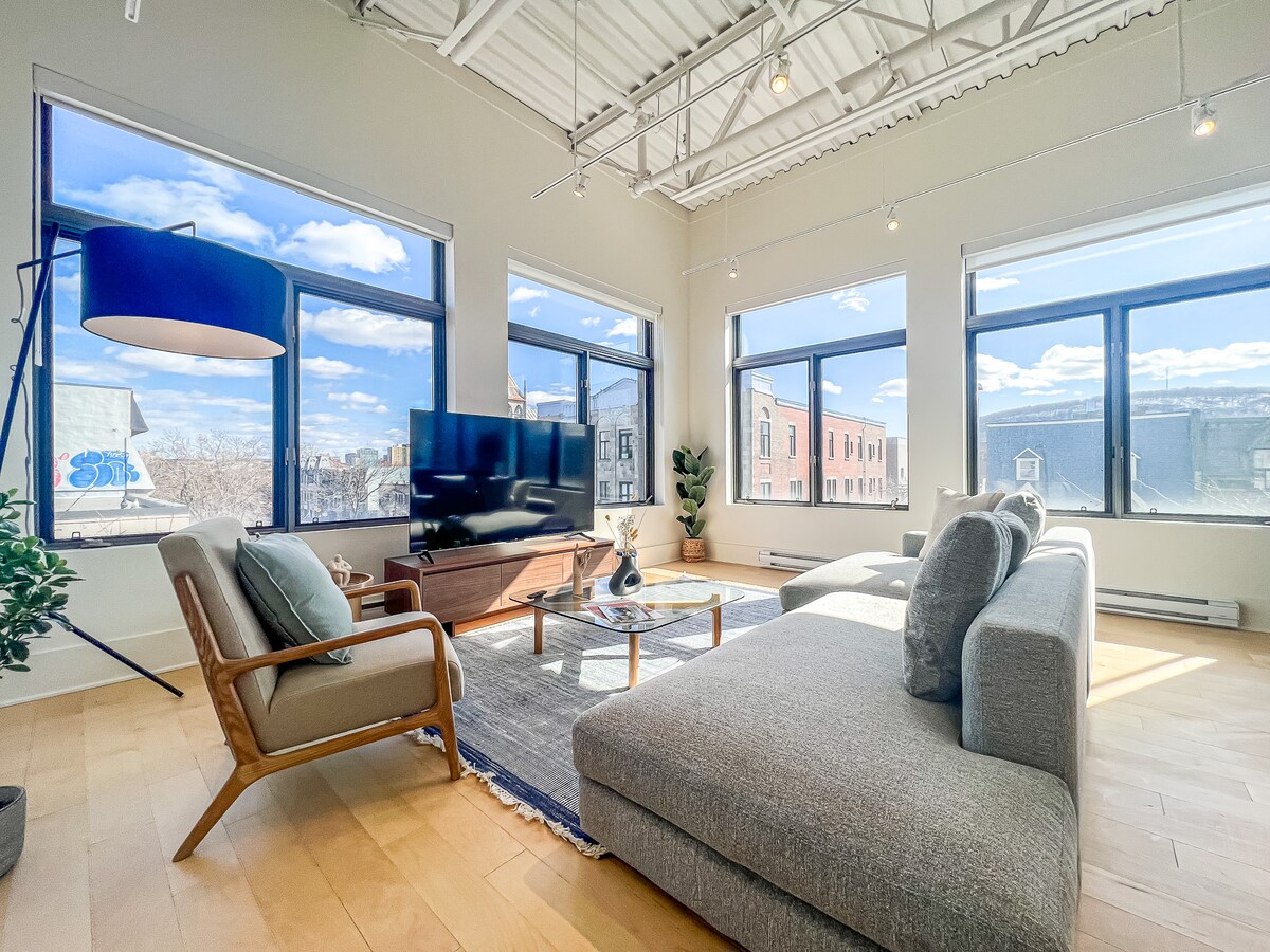 A spacious living area is highlighted by large windows allowing natural light to fill the room. A comfortable sectional sofa is positioned facing a sleek 4k Smart TV. Decorative plants and a stylish coffee table enhance the modern design, set against the backdrop of picturesque city views.