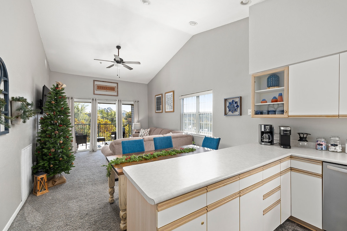 An open living area features a large sectional sofa, with decorative pillows enhancing comfort. Natural light fills the space through floor-to-ceiling windows, leading to a private deck. A festive tree adds a seasonal touch, and a fully equipped kitchen is visible in the foreground.