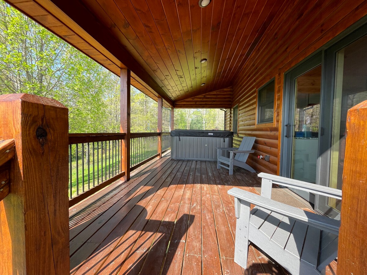 A spacious deck features wooden flooring and log walls, illuminated by natural light. A hot tub is positioned to the side, surrounded by greenery. Two wooden chairs with arms are placed for seating, enhancing the inviting outdoor space.