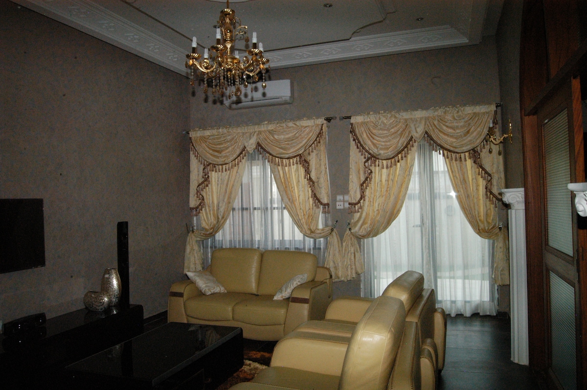 A spacious living area features creamy leather seating arranged around a dark coffee table. Elegant drapes in soft gold tones frame large windows, allowing natural light to illuminate the room. A decorative chandelier hangs from the ceiling, adding a touch of sophistication.
