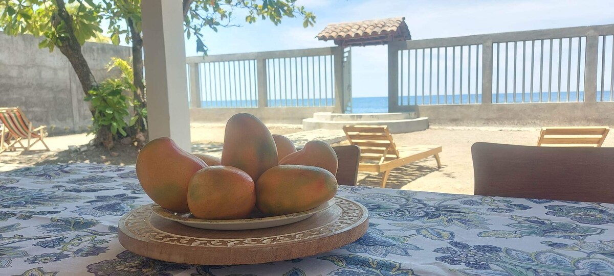 A table adorned with a patterned tablecloth displays a plate of fresh mangoes. In the background, beach loungers are positioned near a view of the Pacific Ocean, framed by a decorative fence and a shaded gazebo.